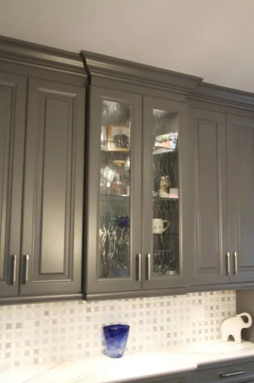 Gray kitchen wall cabinets with some glass-front doors displaying glassware and decorative items, white tiled backsplash, white countertop with a blue glass cup and a white elephant-shaped kitchen accessory.