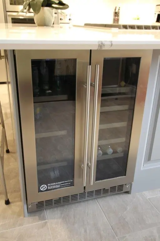 Built-in stainless steel double-door wine cooler with glass panels, under a white countertop in a modern kitchen.