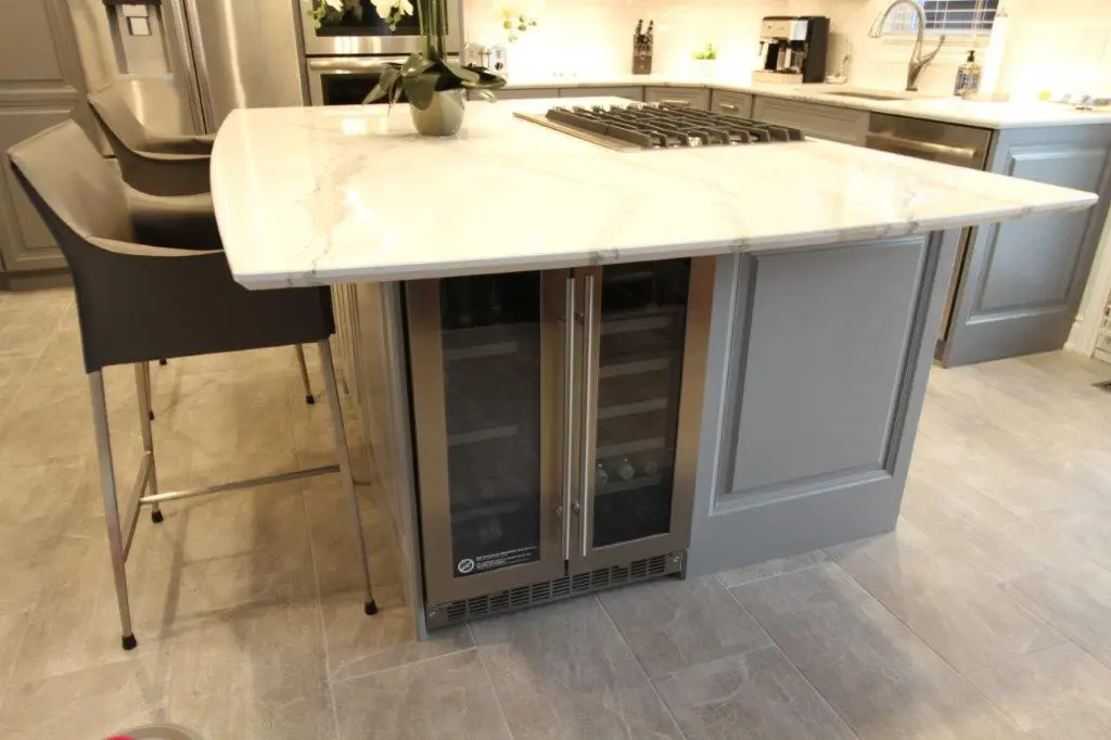 Modern kitchen island with a white marble countertop, built-in wine cooler with glass doors, two gray bar stools, and a gas stovetop, all set on a light gray tiled floor.