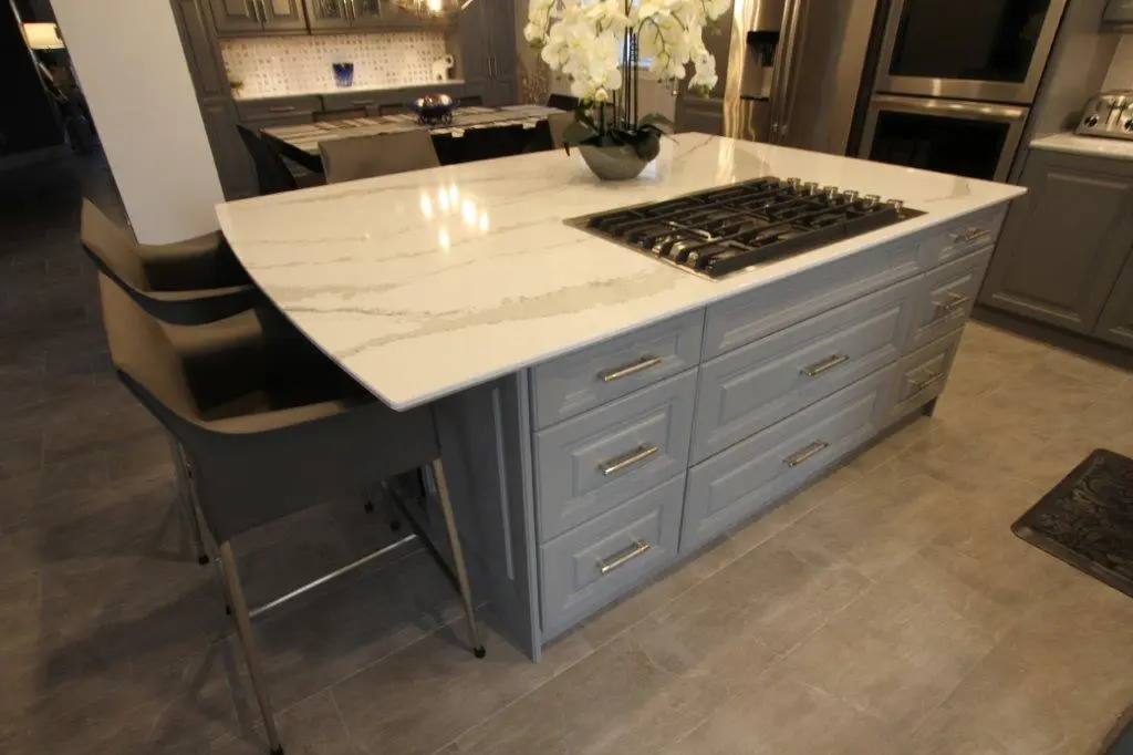 Modern kitchen island with a white marble countertop, built-in gas stove, gray cabinets with silver handles, two gray chairs tucked underneath on the left, and a potted orchid on top.