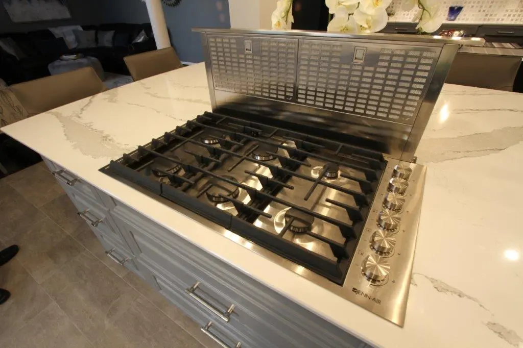 Modern built-in stainless steel cooktop with six burners and control knobs on the right side, installed in a white marble countertop with gray veining, set in a kitchen island with gray cabinetry and stainless steel handles.