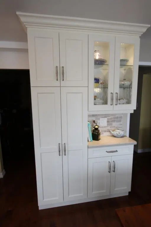 White kitchen cabinetry with multiple doors and metal handles, a countertop with decorative items including a jar and a ceramic dish, and a glass-front upper cabinet displaying dishes, set against a tiled backsplash and dark wood flooring.