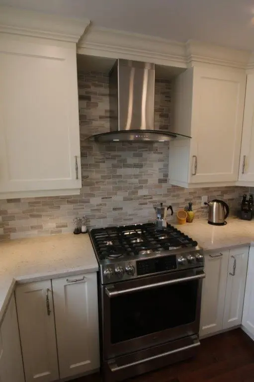 Modern kitchen with a stainless steel gas stove and oven, white cabinetry, a beige and gray tiled backsplash, and a stainless steel range hood above the stove.
