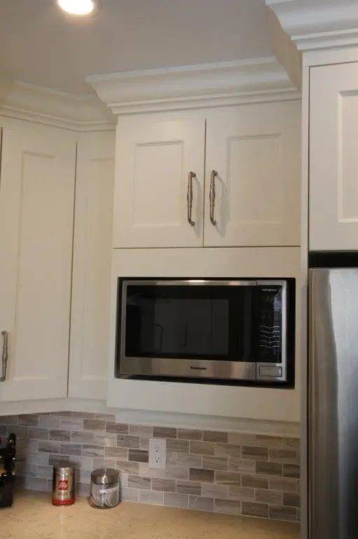 Built-in microwave oven with stainless steel trim installed in white kitchen cabinetry above a tiled backsplash featuring neutral tones, with a stainless steel refrigerator partially visible on the right and kitchen counter with items below the backsplash.