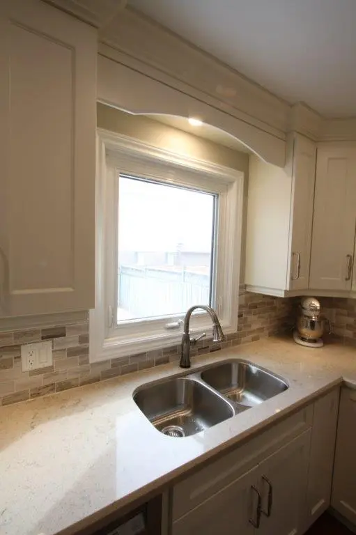 Modern kitchen countertop with a double stainless steel sink under a window, beige cabinets, a tiled backsplash, and a mixer on the corner of the counter.