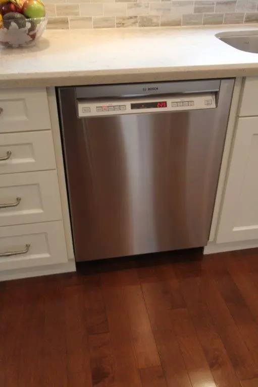 Stainless steel Bosch dishwasher integrated into white kitchen cabinetry with a beige countertop and wooden floor.