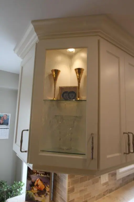 Cream-colored kitchen cabinet with a glass display section illuminated from above, containing two gold decorative goblets and a small dish on the top shelf and a clear glass vase on the bottom shelf, with part of a calendar and a cookbook visible in the background.