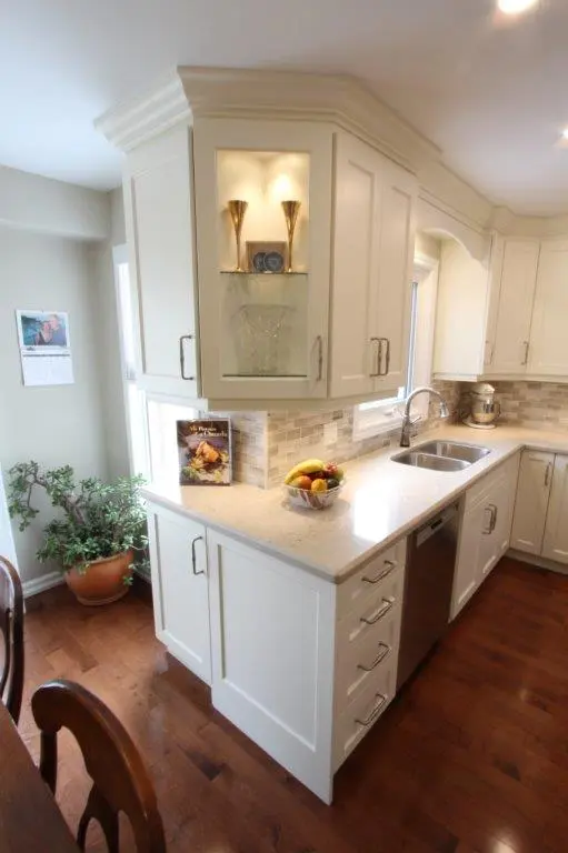 Modern kitchen corner with white cabinetry, granite countertop, stainless steel sink and dishwasher, bowl of fruit, a cookbook, a potted plant near the corner, and wood flooring.