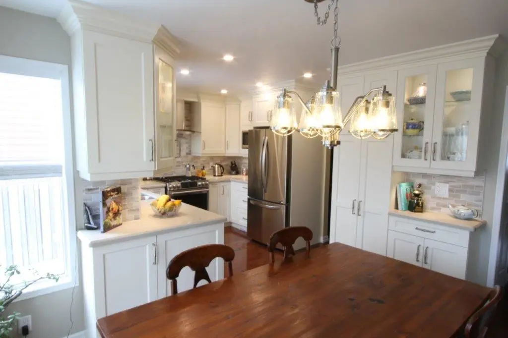 Bright kitchen with white cabinets, stainless steel refrigerator and stove, wooden dining table with three wooden chairs, and a hanging light fixture with multiple bulbs
