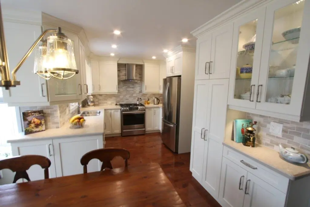 Modern kitchen with white cabinetry, stainless steel refrigerator and stove, a glass-front cabinet with dishes, wooden dining table with chairs, and pendant light fixture.