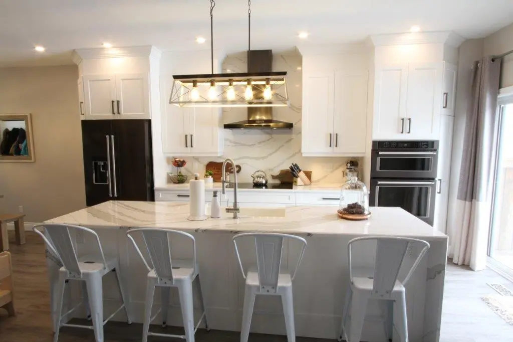 Modern kitchen with a large white marble island featuring four metal bar stools, pendant light fixture, white cabinets, black refrigerator, double wall ovens, and a glass door with curtains to the right.