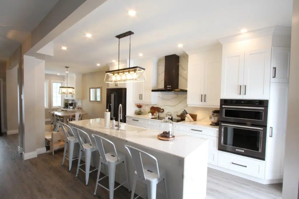 Bright modern kitchen featuring a large white island with metal bar stools, white cabinetry, a black refrigerator, double wall ovens, and pendant lighting above the island, with a dining area visible in the background.