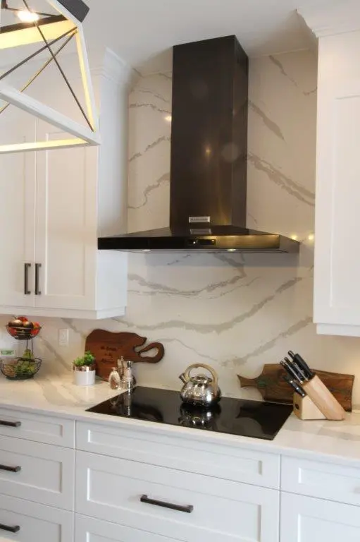 Modern kitchen countertop with white cabinetry, a black electric stove with a stainless steel kettle, a black range hood, wooden cutting boards, a knife block, and various small kitchen decor items on a marble-patterned backsplash.
