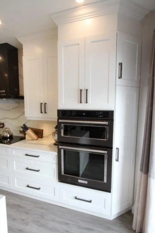 Modern kitchen interior featuring white cabinetry with black handles, built-in double oven, and a countertop with a kettle and knife block, set against a marble-patterned backsplash and light gray wood flooring.
