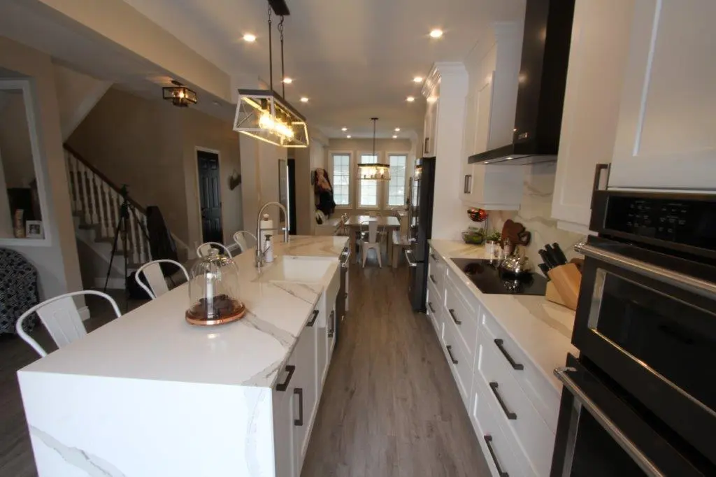 Modern kitchen with white marble countertops, white cabinets, black handles, and a central island with bar stools, illuminated by pendant lights and recessed ceiling lights, leading to a dining area with a rectangular table and chairs in the background.