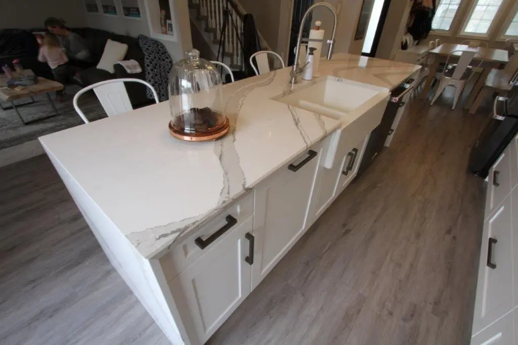 Modern kitchen island with white marble countertop featuring gray veining, undermount sink, and white cabinetry with dark handles, surrounded by white metal chairs and wood-look flooring.