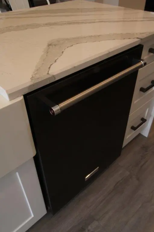 Black built-in dishwasher with a stainless steel handle installed under a white and gray marbled countertop in a kitchen with white cabinetry and gray wood flooring