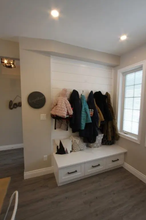 Entryway with a white bench featuring three drawers, cushioned with decorative pillows, and a wall-mounted coat rack holding several winter jackets beneath recessed lighting and near a window with blinds.