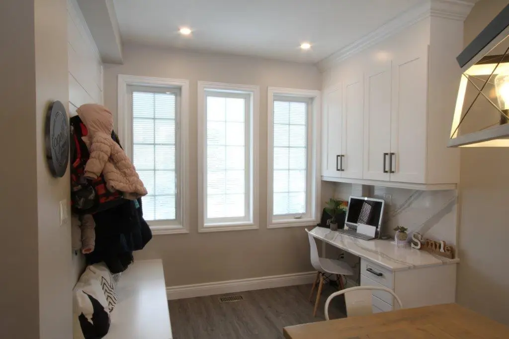 Bright home office corner with three tall windows, a white desk with a computer and plants, white upper cabinets, a white chair, and a coat rack with jackets and hats next to a bench with pillows.