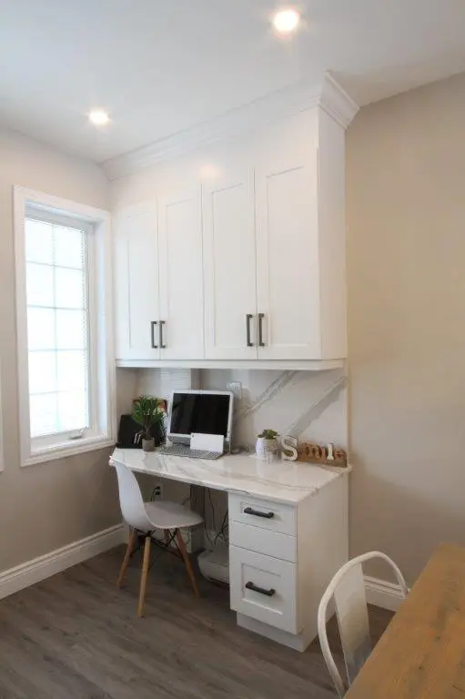 Small home office nook with white cabinetry, marble-patterned desk, computer, potted plants, and a white chair, next to a window with blinds and gray wood flooring.