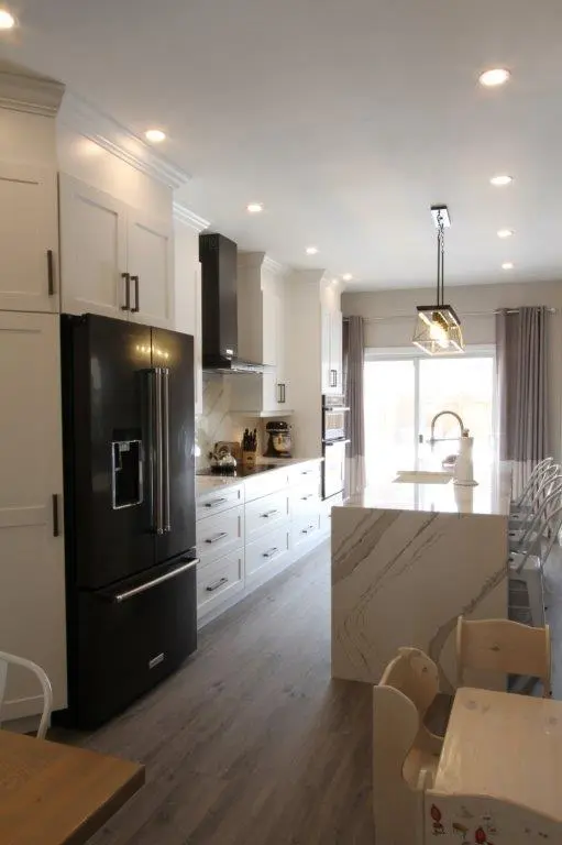 Modern kitchen with white cabinetry, a black refrigerator, marble island with a sink, and metal bar stools, featuring recessed lighting and a large window with curtains.