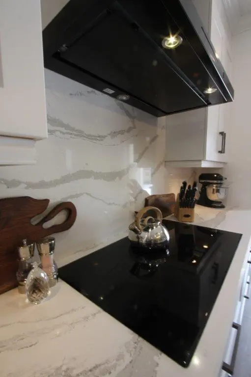 Modern kitchen countertop with a black electric stovetop, stainless steel kettle, spice containers, wooden cutting board, knife block, and a black range hood overhead.