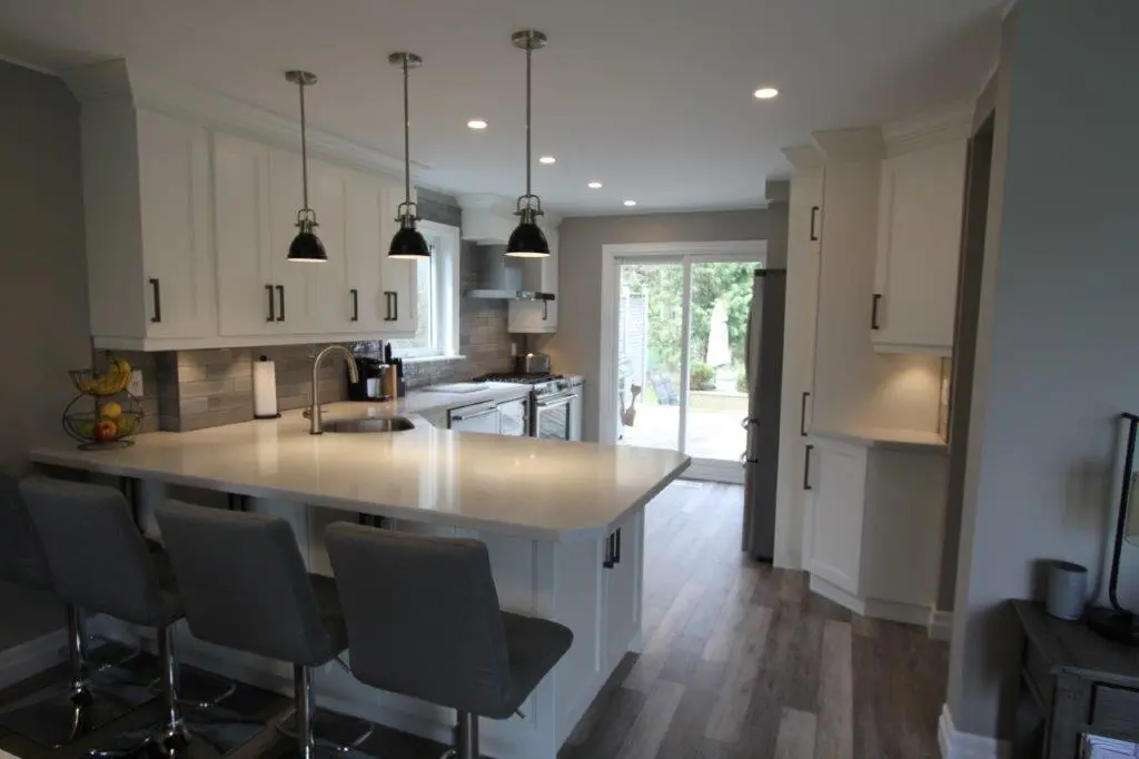 Modern kitchen with white cabinets, gray backsplash, three hanging pendant lights, a large white island with four gray bar stools, stainless steel appliances, and sliding glass doors leading to an outdoor area with greenery.