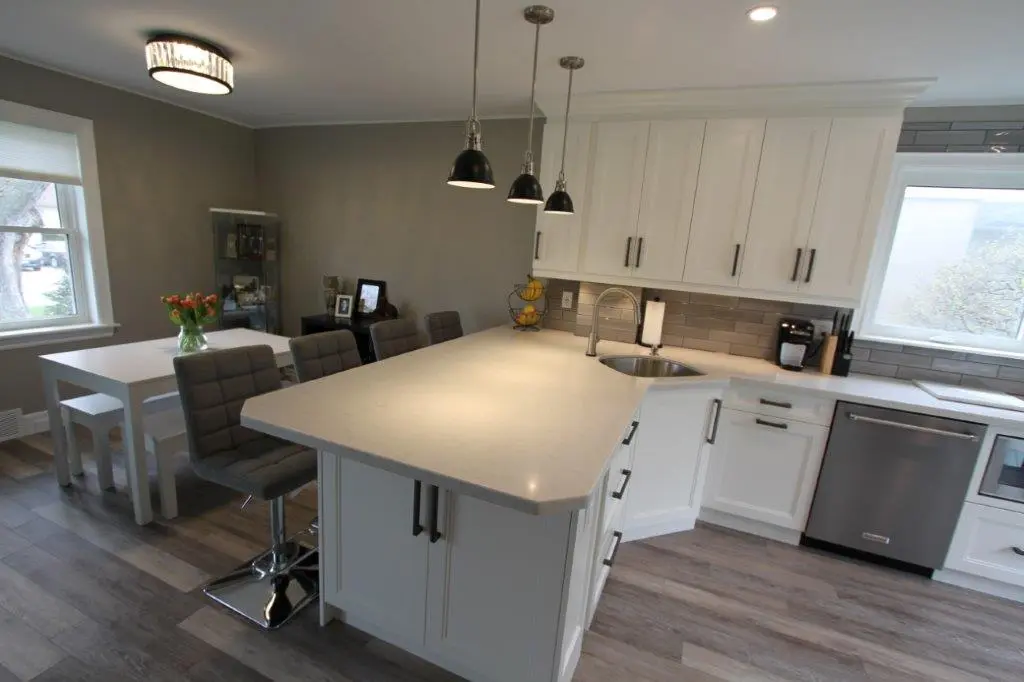 Modern kitchen with white cabinets, a large light gray island with built-in sink, three black pendant lights, gray padded bar stools, stainless steel dishwasher, and a dining table with a vase of orange flowers near a window