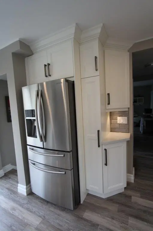 Modern kitchen corner featuring a stainless steel Samsung French door refrigerator with two lower freezer drawers, surrounded by white cabinetry with black handles and a small illuminated countertop with a tiled backsplash and electrical outlets.