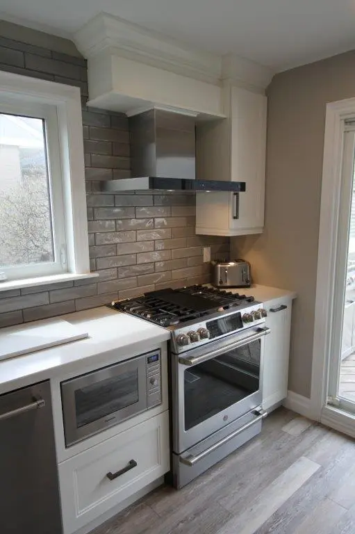 Modern kitchen corner featuring a stainless steel gas stove with oven, a microwave built into white cabinetry, a stainless steel range hood, gray subway tile backsplash, a toaster on the white countertop, and a sliding glass door on the right.