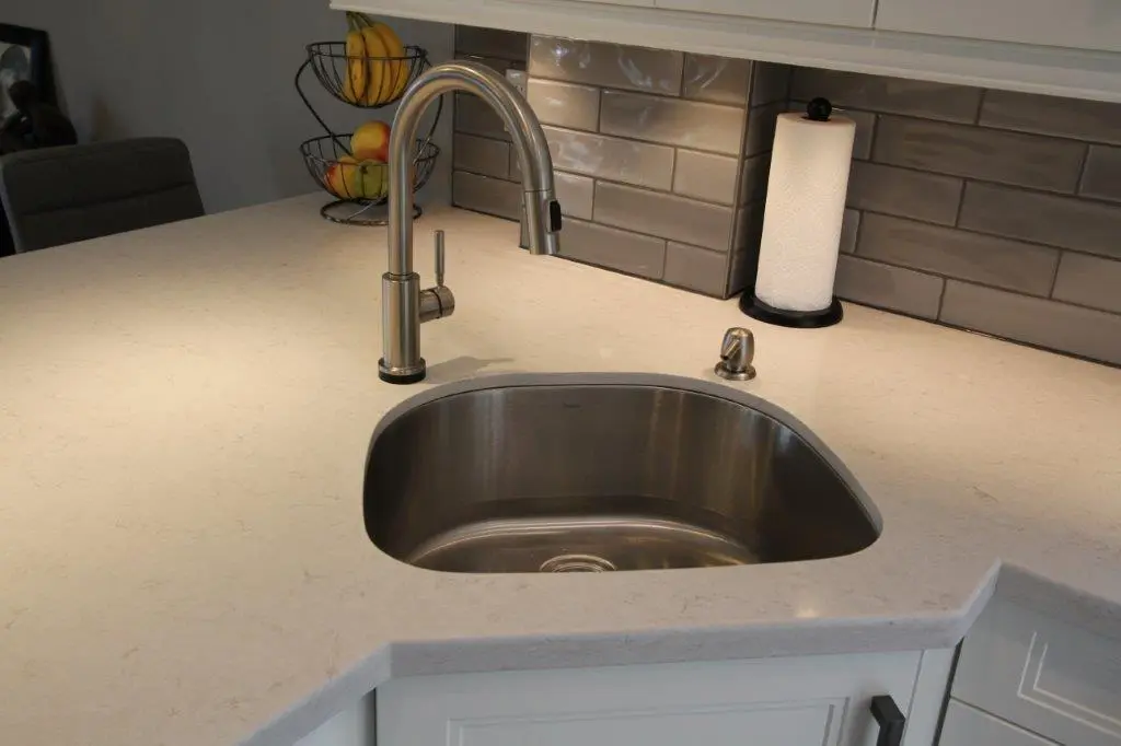 Modern kitchen countertop with an undermount stainless steel sink, a tall curved faucet, a soap dispenser, and a paper towel holder against a gray subway tile backsplash and white cabinetry
