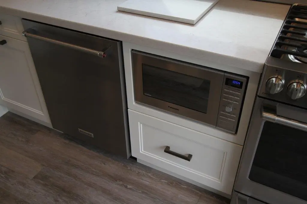 Modern kitchen setup featuring a stainless steel microwave built into white cabinetry, adjacent to a stainless steel dishwasher on the left and a gas stove on the right, with a white countertop above.