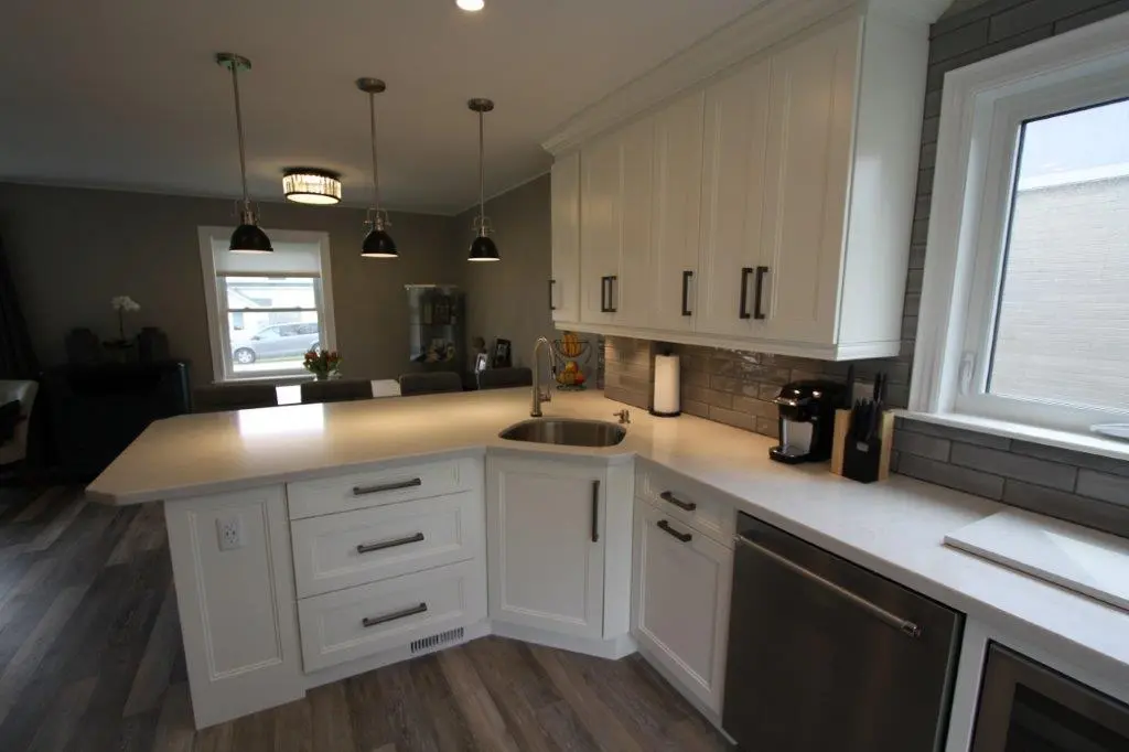 Modern kitchen with white cabinetry, gray subway tile backsplash, and a large white countertop island featuring a built-in sink, with pendant lights hanging above and a stainless steel dishwasher beneath the counter near a window.