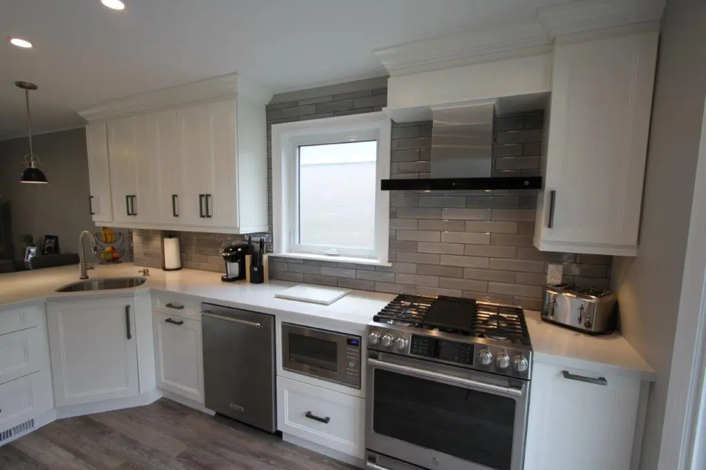 Modern kitchen corner with white cabinetry, stainless steel appliances including a dishwasher, microwave, gas stove with oven, and a toaster, gray subway tile backsplash, a window above the countertop, and a sink with a curved faucet.