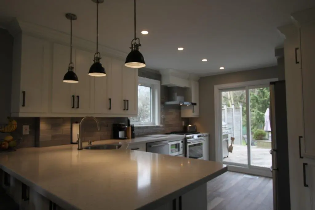 Modern kitchen with white cabinets, a large island with a smooth countertop, three black pendant lights hanging above, stainless steel appliances, a window above the sink, and a sliding glass door leading to an outdoor patio.