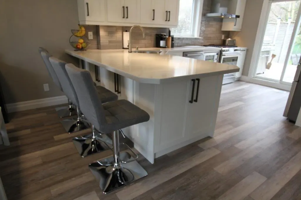 Modern kitchen with a white island countertop featuring three gray cushioned bar stools with chrome bases, wooden flooring, and stainless steel appliances near a glass sliding door.