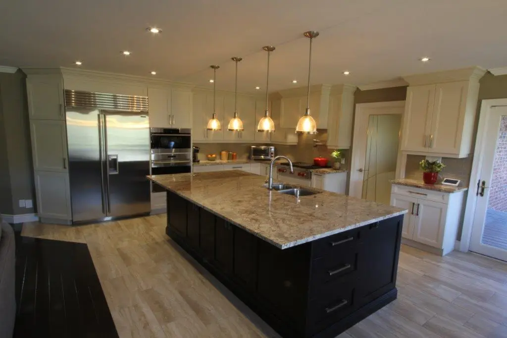 Modern kitchen with a large granite island countertop featuring a double sink, surrounded by white cabinetry, stainless steel refrigerator and built-in oven, pendant lights hanging above the island, light wood flooring, and glass door leading outside.