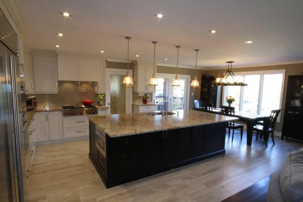 Modern kitchen with a large central island featuring a granite countertop, hanging pendant lights above, white cabinetry, stainless steel appliances, and a dining area with a table and chairs near large windows.