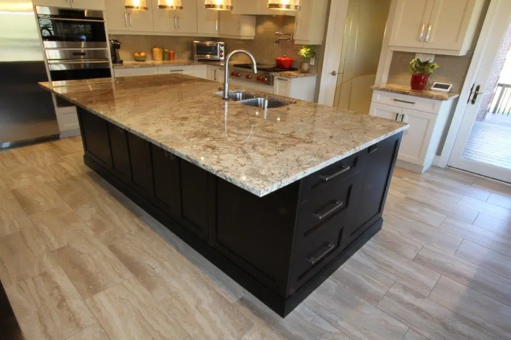 Modern kitchen with a large granite island countertop featuring a built-in sink and dark cabinetry, surrounded by light-colored tile flooring and white cabinets in the background.