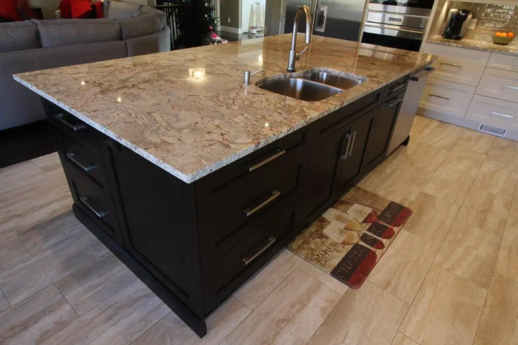 Kitchen island with a polished granite countertop and built-in double stainless steel sink, dark wood cabinets below, on a tiled floor with a small mat featuring images of drinks in front of the sink area.