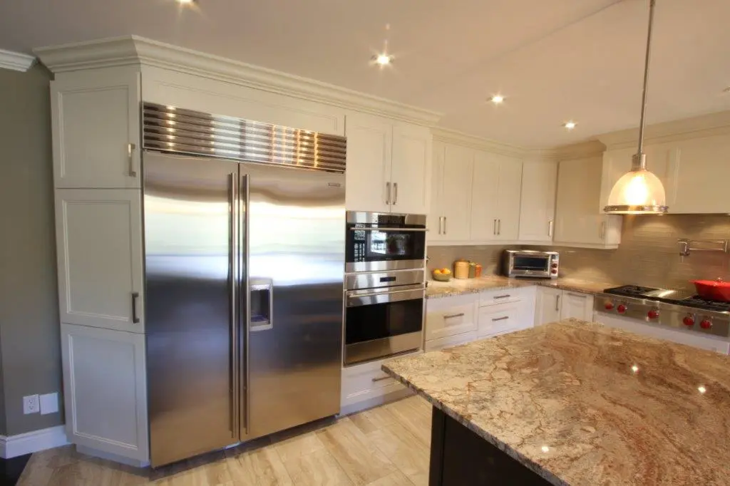 Modern kitchen with stainless steel double-door refrigerator, built-in microwave and oven, white cabinetry, granite countertop island, and pendant lighting over the island.