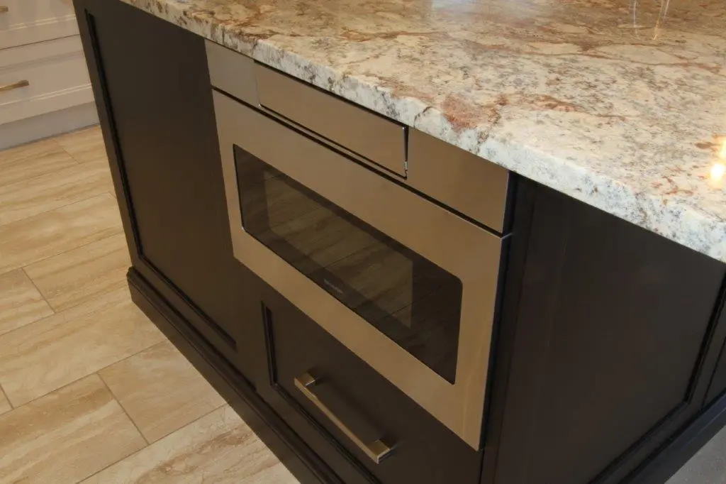 Built-in stainless steel microwave installed in a dark wooden kitchen island with a beige and brown granite countertop and light wood tile flooring.