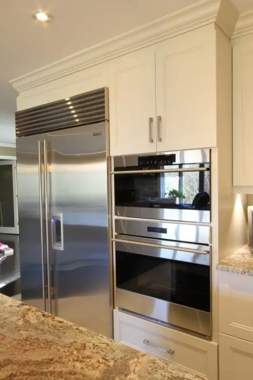 Stainless steel side-by-side refrigerator next to built-in double oven with white cabinetry and granite countertop in modern kitchen
