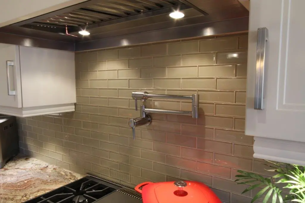 Modern kitchen backsplash with beige subway tiles, a stainless steel pot filler faucet mounted on the wall, white cabinets with metal handles, a granite countertop, part of a gas stove, and a red cast iron pot.
