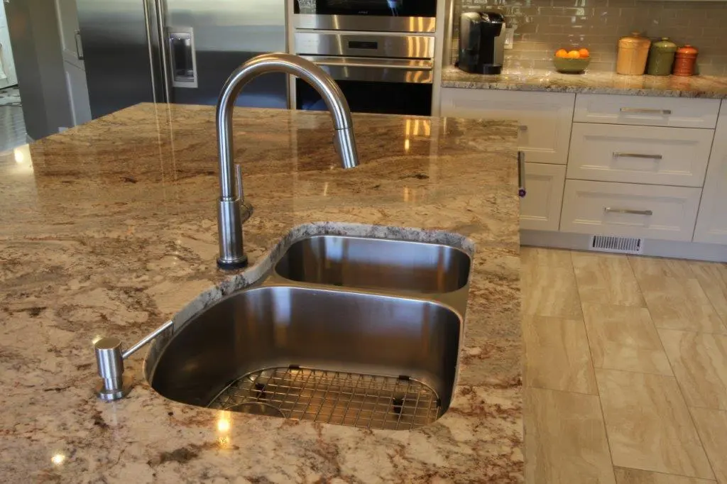 Modern kitchen countertop with a double stainless steel sink and a tall curved faucet, surrounded by beige and brown granite, with white cabinets and stainless steel appliances in the background