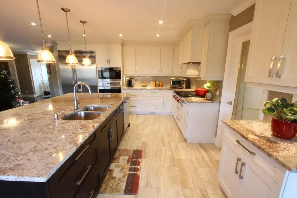 Modern kitchen with a large granite island featuring a double sink and pendant lights, light-colored cabinets and countertops, stainless steel appliances including a refrigerator and oven, and a light wooden floor with a decorative rug near the island.