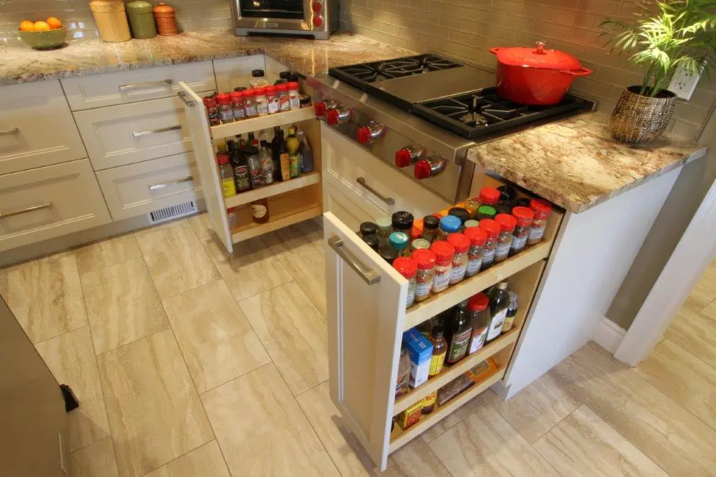Modern kitchen with beige tiled floor, granite countertops, and two open pull-out spice racks filled with bottles and jars next to a stainless steel gas stove with red knobs and a red pot on top, and a potted plant on the counter.