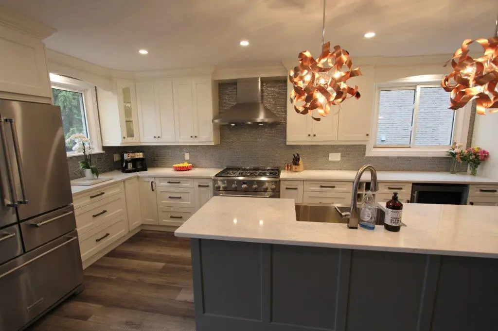 Modern kitchen with white upper and lower cabinets, stainless steel refrigerator and stove, gray island with white countertop and sink, decorative pendant lights, and wood-look flooring.