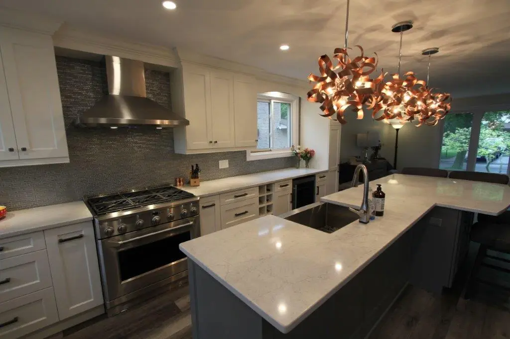 Modern kitchen with white cabinetry, a gray stone countertop island with a built-in sink and faucet, stainless steel stove with hood, and artistic pendant lighting above the island, with a window and flowers near the back counter and large windows showing greenery outside.