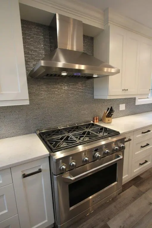 Modern kitchen featuring a stainless steel gas stove with multiple burners, a matching stainless steel range hood above, white cabinetry, a gray tiled backsplash, and a white countertop with a knife block and seasoning shakers.